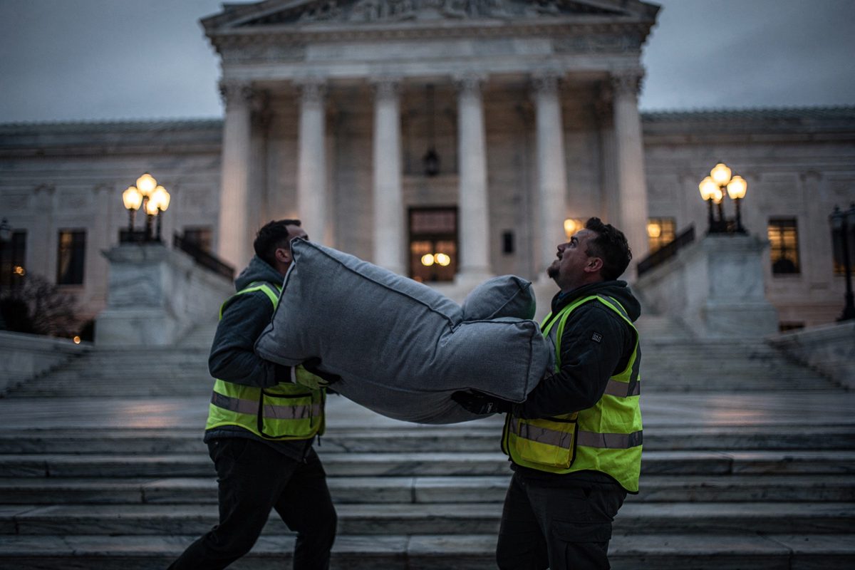 Wide shot at dusk as streetlamps flicker on, two delivery workers in reflective vests carrying a large grey beanbag chair up the broad stone steps of a.