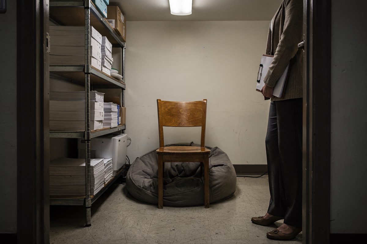 Mid shot under a single bare ceiling bulb inside a windowless supply closet, a metal shelving unit still stacked with reams of printer paper along one wall.
