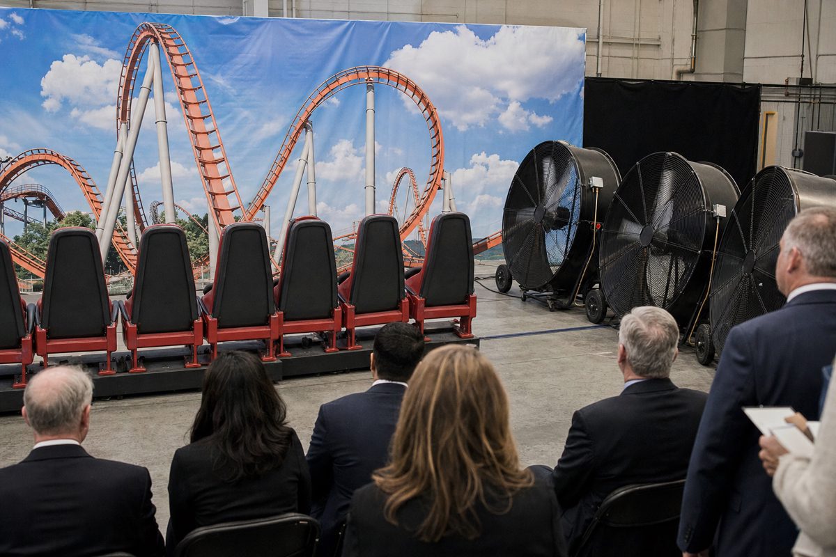 Realistic press photograph inside a government demonstration hall showing a row of stationary amusement-ride seats facing large industrial fans and a painted.