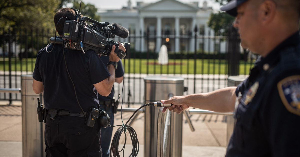Mid-shot in bright late-morning sun at a White House pedestrian gate on Pennsylvania Avenue, a television crew seen from behind on the pavement with a.