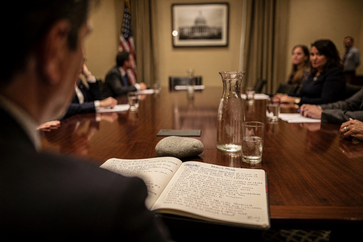 Over-the-shoulder view in warm lamplight inside a hotel meeting room, a facilitator's open notebook covered in handwritten bullet points while a smooth grey.