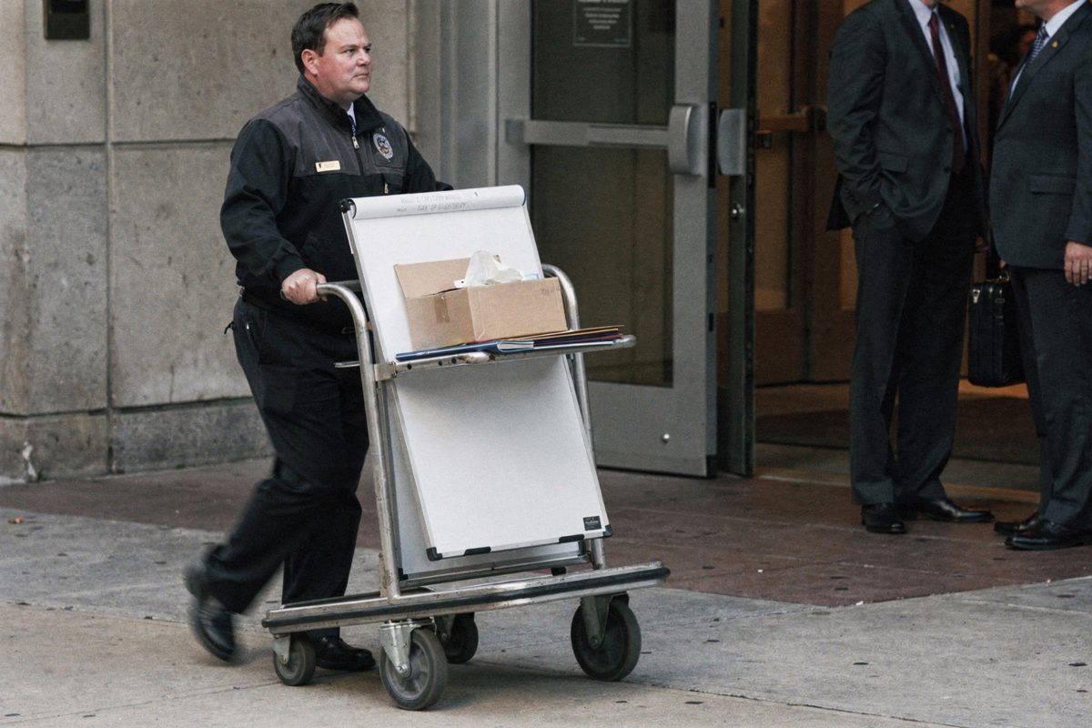 Mid-shot in flat late-afternoon light on a Washington sidewalk outside a hotel service entrance, a porter wheeling a luggage trolley loaded with a flip-chart.