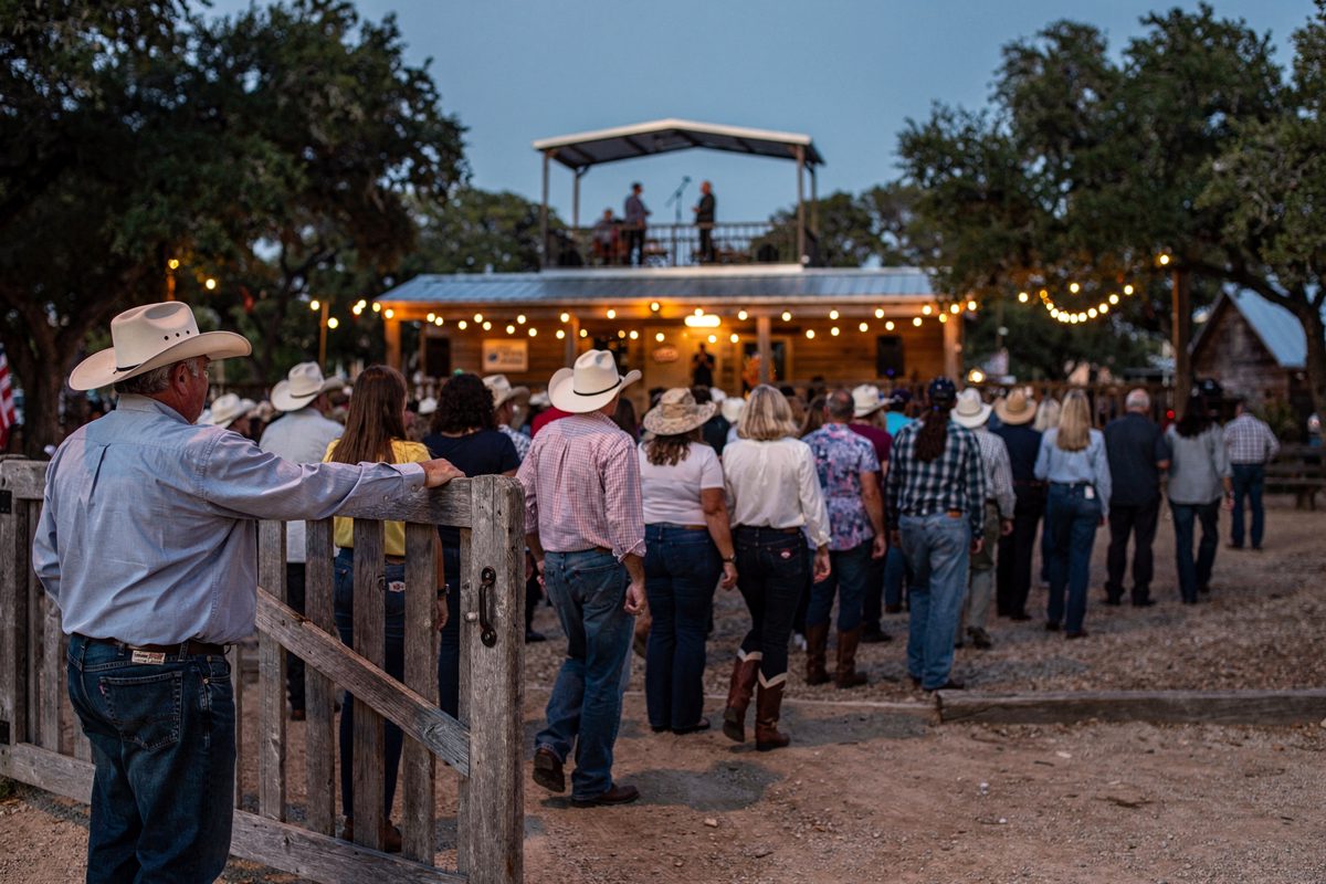 Wide shot from the back of an outdoor Hill Country dance hall rally at dusk, a crowd of supporters seen from behind facing a distant stage while one man near.