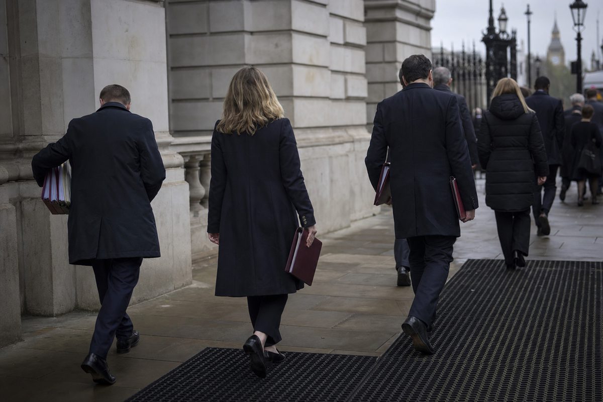 A realistic press photograph of anonymous officials leaving a government building carrying document folders under overcast London skies, faces turned away and.