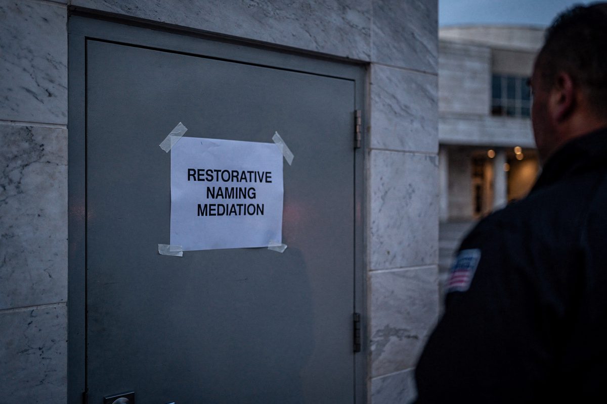 Close shot at dusk of a paper notice reading "RESTORATIVE NAMING MEDIATION" taped to a grey service door on a marble performing arts complex, a courier's.