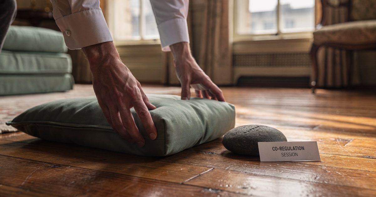 Tight morning light across a historic riverside parlor floor, a staffer's hands cropped at the cuffs lowering a sage floor cushion beside a dark river stone.