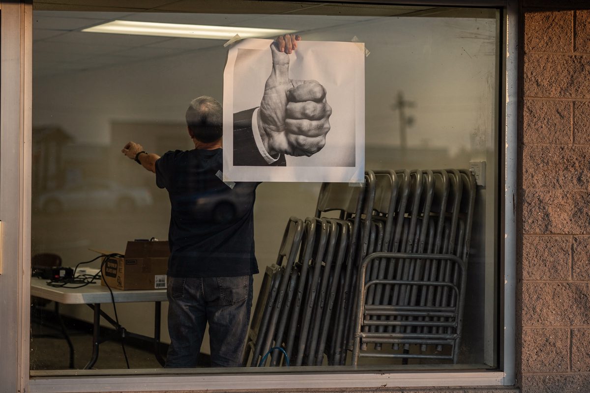Wide late-afternoon shot from the sidewalk into a strip-mall campaign office window, two volunteers with their backs turned taping up an enlarged.