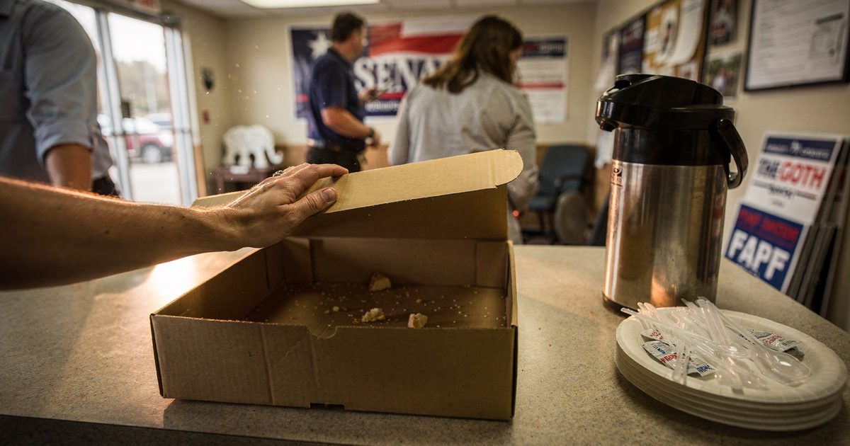Close detail in bright early-morning sun at the breakfast counter of a strip-mall Senate campaign office, a hand cropped at the wrist lifting the lid of an.