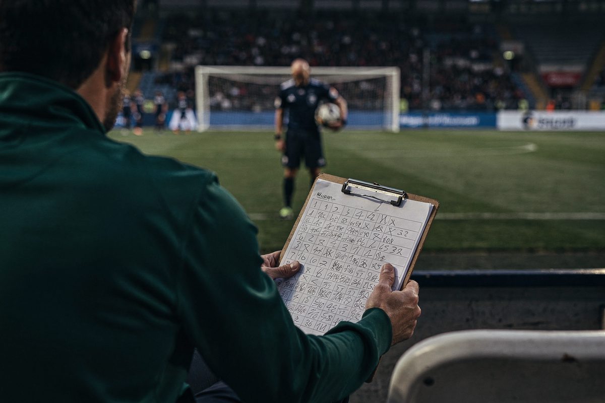 Shot from directly behind a dugout bench at dusk, a coaching staff member in a green tracksuit seen from the shoulders down holding a clipboard covered in.