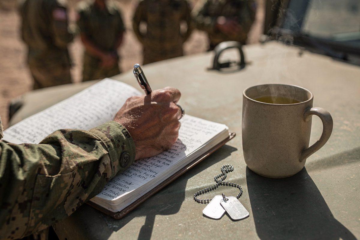 Close-up in harsh midday sun of a camouflage-sleeved hand writing in a spiral notebook propped on the hood of a parked Humvee, a set of dog tags pooled beside.