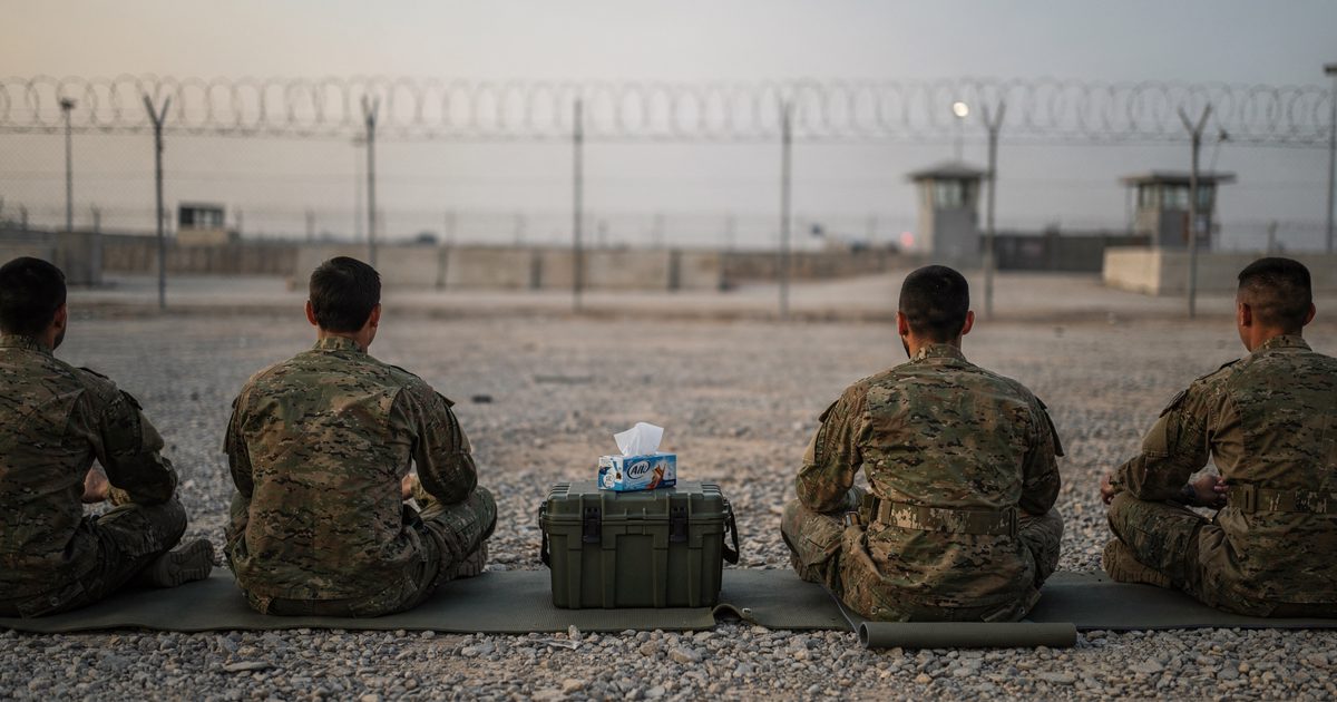 Soldiers in camouflage sit cross-legged on yoga mats laid over gravel beside a razor-wire border fence at dawn, photographed from behind, a tissue box resting.
