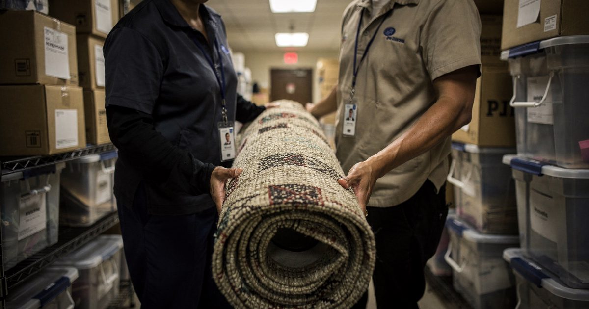 Harsh overhead light in a municipal storage corridor as two election workers cropped from the neck down struggle to carry a rolled-up woven therapy rug past.