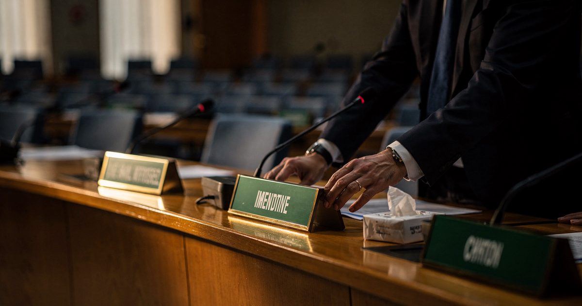 Late-afternoon sun slanting across a long curved delegates' desk in a vast assembly hall, an aide cropped at the elbows sliding a green country nameplate.