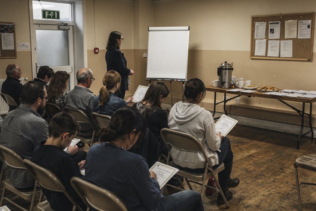 Wide shot from the back of a strip-lit village hall, rows of stacking chairs half-filled with people seen from behind facing a flipchart easel, a tea urn.