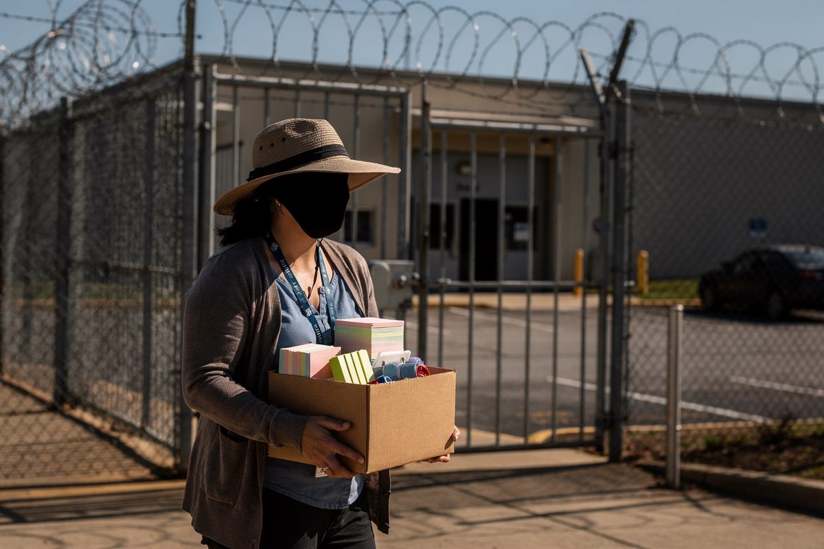Medium shot in hard bright morning sun at the razor-wire perimeter gate of a Nashville maximum security prison, a woman in a cardigan with a lanyard carrying a.