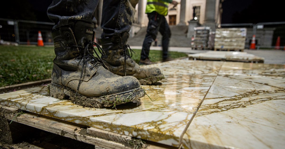A pair of mud-caked steel-toed work boots resting heavily on a pallet of pristine, uninstalled gold-veined Italian marble under the harsh glare of temporary.