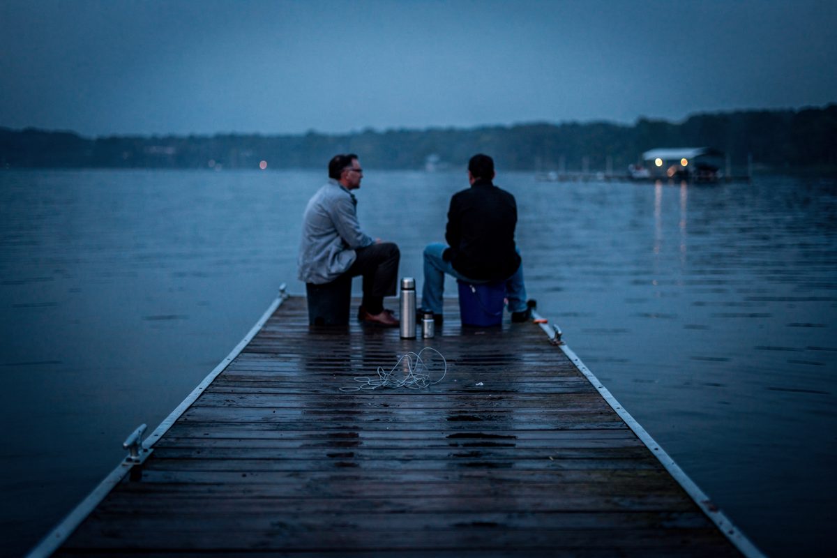 In blue hour at the far end of a wooden fishing pier, two small figures sit with a thermos and a coil of line between them, faces unreadable at that distance.