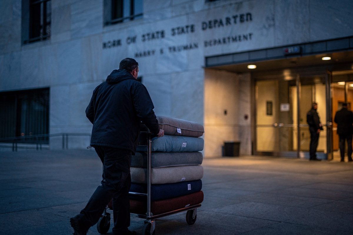Wide shot at blue hour outside the State Department's Harry S Truman building in Washington, a courier seen from behind wheeling a hand truck stacked with.