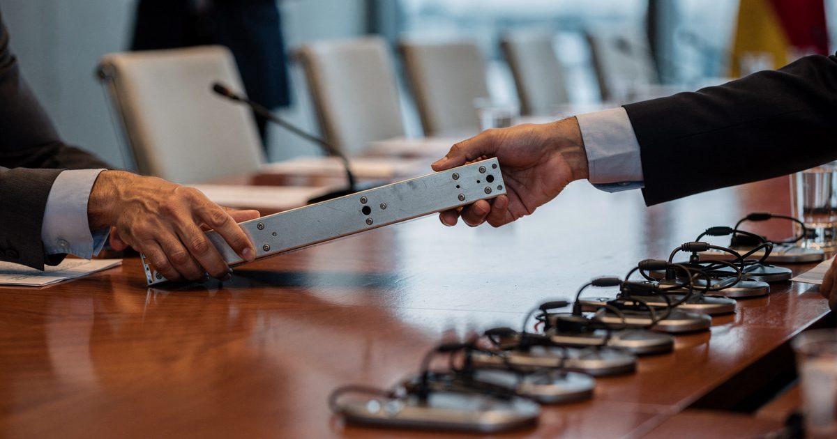 Close overhead shot in cool mid-morning light along a polished Geneva conference table, two delegates' hands cropped at the cuffs passing a short metal rail.