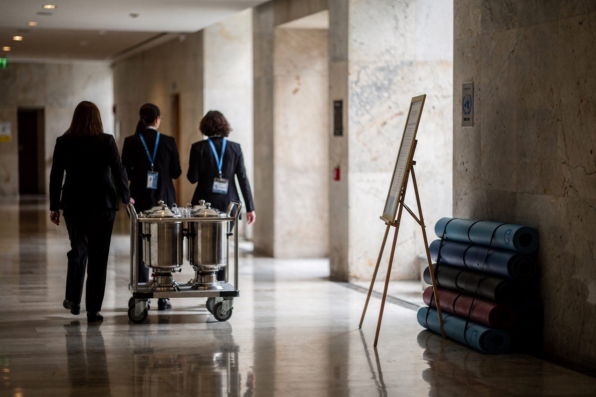 Wide late-afternoon shot of a marble United Nations annex corridor, three lanyarded staff seen from behind wheeling a stainless tea urn past a folding easel.