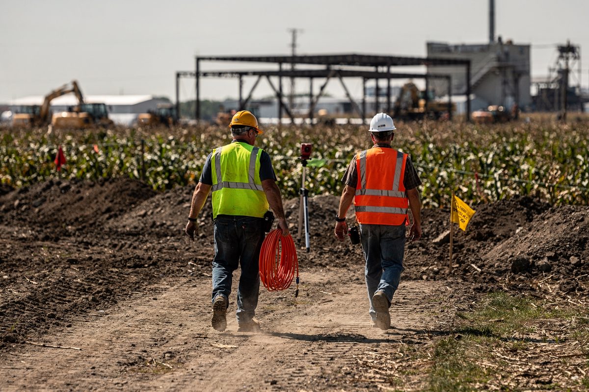 Medium shot under a hard noon sky at the rim of a cornfield, two surveyors seen only from the back walking a straight line across fresh grading, one carrying a.