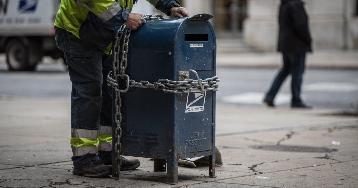 Pale morning light on a cracked concrete sidewalk as a municipal worker in high-visibility gear wraps thick metal chains around a blue postal collection box.