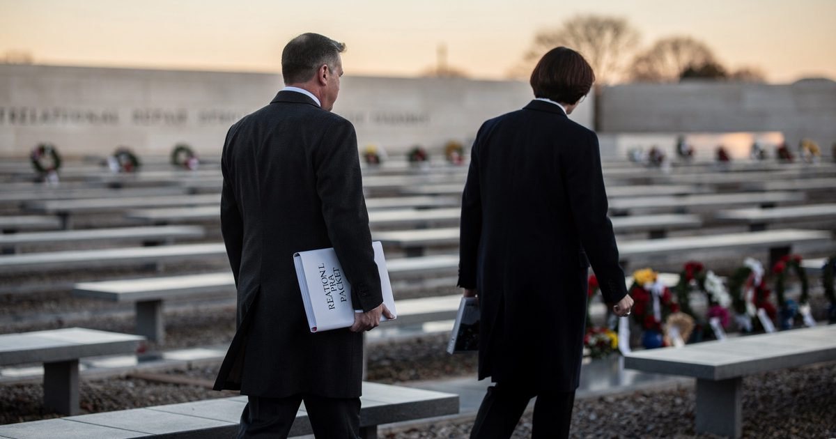 Medium shot at sunrise across the Pentagon memorial grounds, two protocol staff seen from behind in dark coats walking between rows of cantilevered benches as.