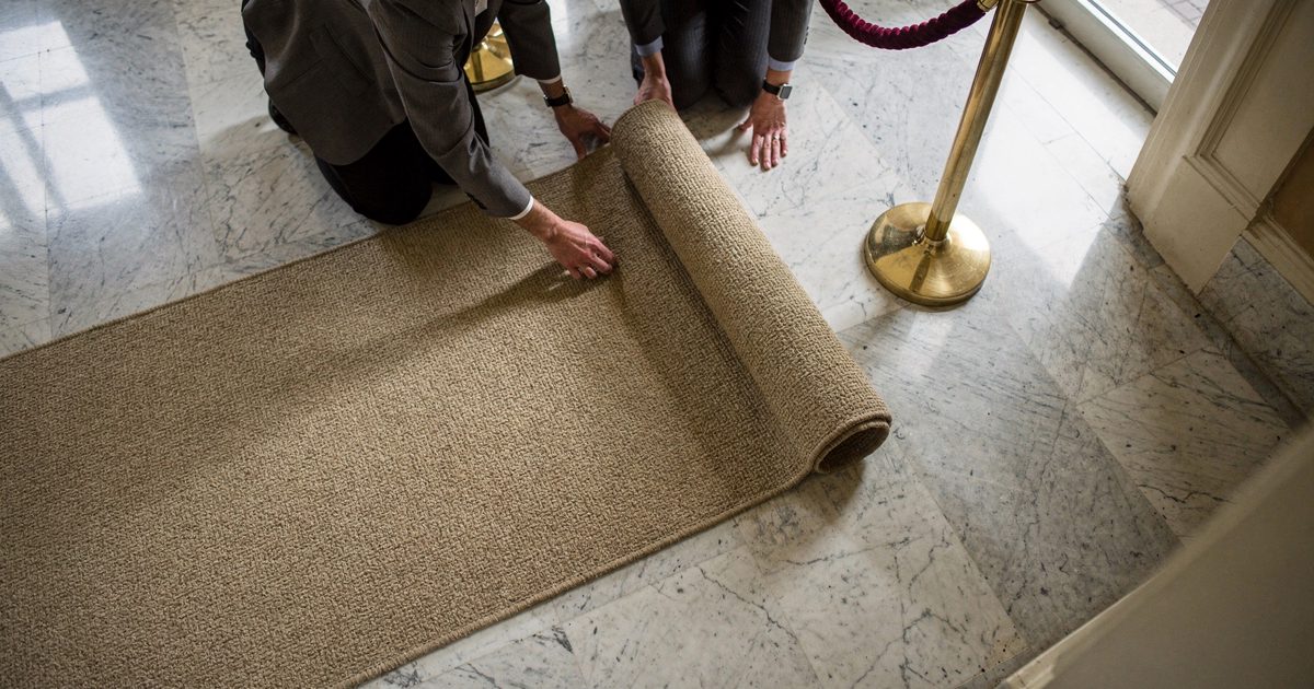 Overhead shot in a sunlit Capitol Hill hallway on a bright morning, two legislative aides seen from the shoulders down rolling a woven jute rug across the.