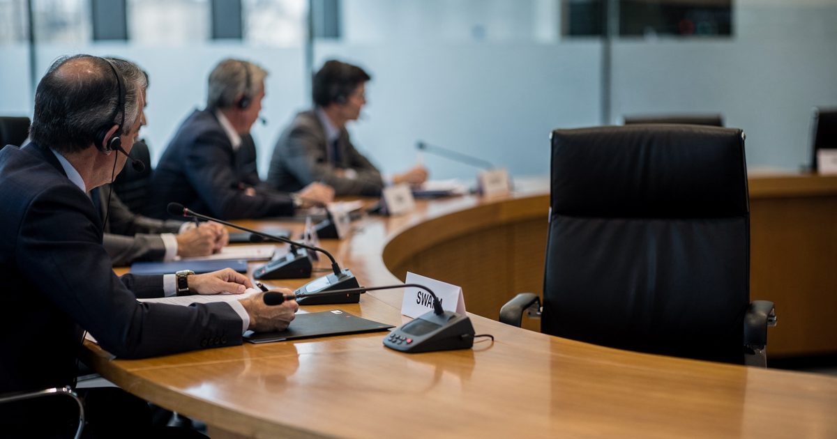 Medium shot in cool mid-morning light along a curved Geneva conference desk, four delegates cropped at the chin turning toward one unoccupied leather chair.