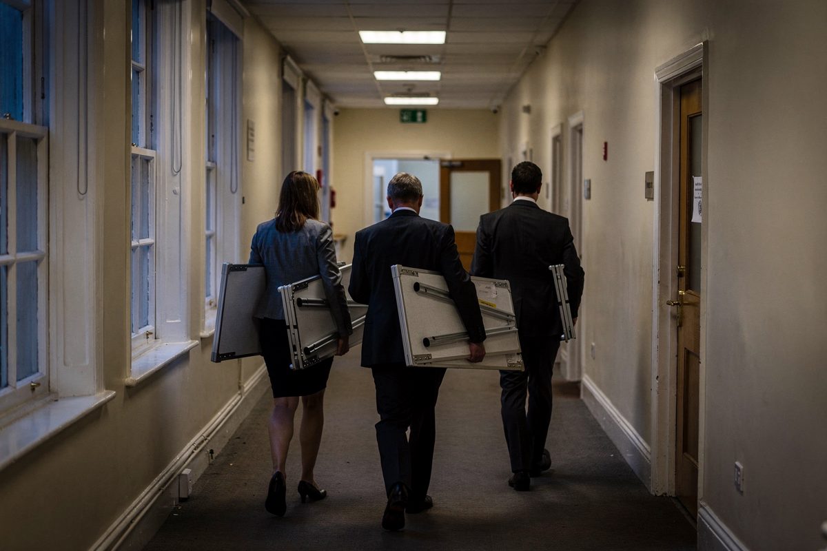 Wide shot down a government corridor at dusk with tall sash windows on one side, three civil servants seen from behind and slightly above carrying folded.