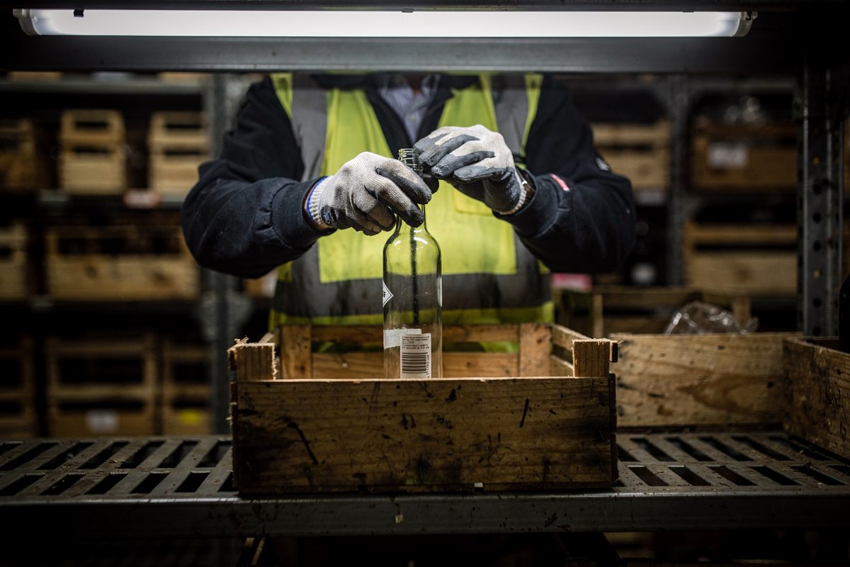 Cold strip lighting in a Leeds drinks wholesaler's stockroom at night, a warehouse worker cropped at the shoulders lifting a single unlabelled glass bottle out.