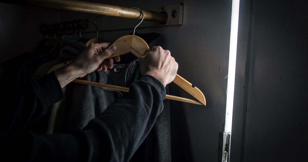 A close-up shot of a person's hands tightly gripping a wooden clothes hanger inside a cramped, dark space, illuminated only by a harsh sliver of fluorescent.