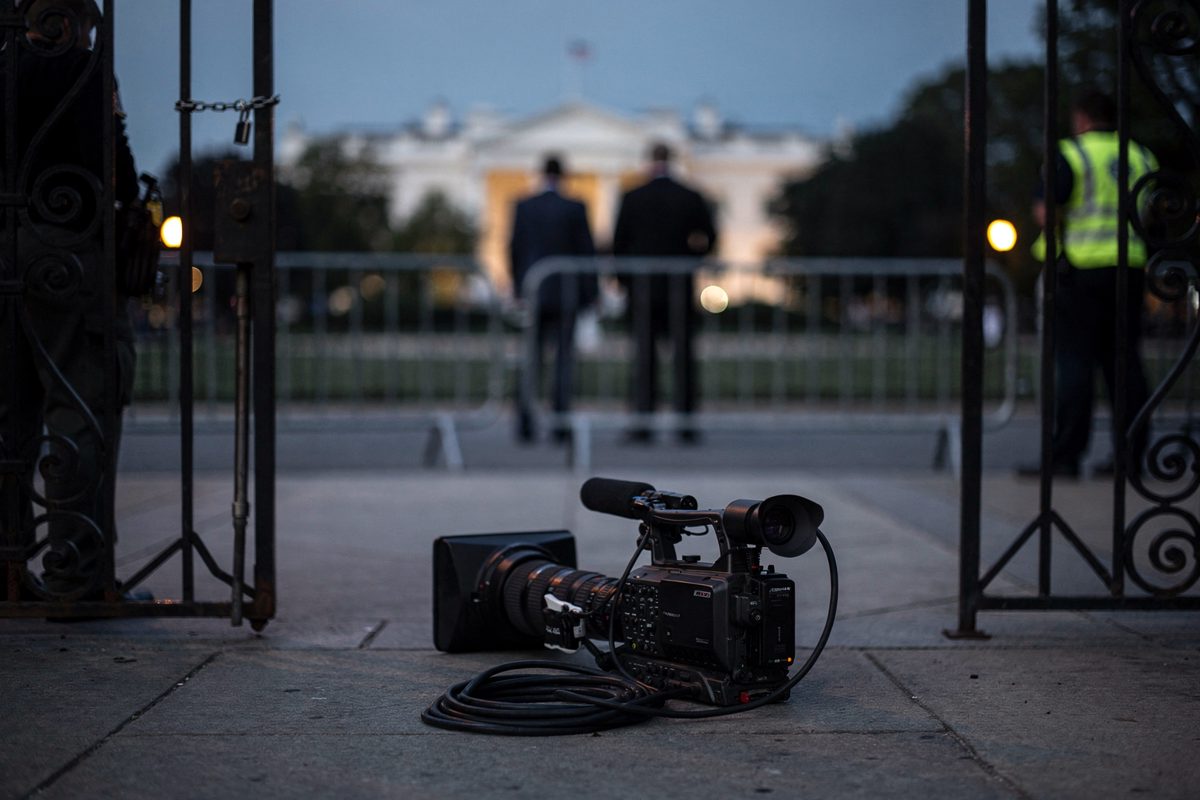 Wide shot through a wrought-iron security gate at dusk, a solitary broadcast camera resting lens-down on the paved sidewalk beside a coiled black cable.