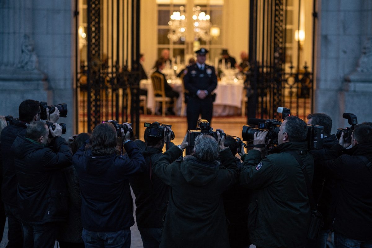 Mid-shot in cool evening light at the closed east gate, a knot of journalists in dark coats with still cameras raised so the cameras hide their faces, a.