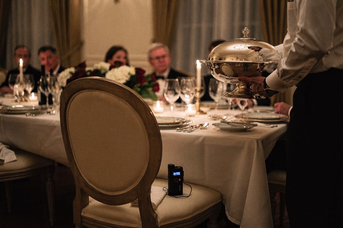Tight candlelit shot across a long state-dinner table of an empty chair pulled slightly out, a small audio recorder with a dark indicator resting on the seat.