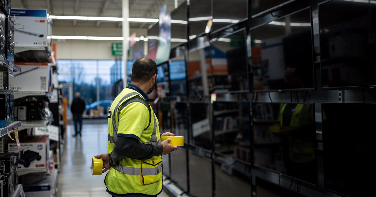 Wide shot from a high angle inside a brightly lit retail electronics aisle just before dawn, a worker in a high-visibility vest standing motionless before a.