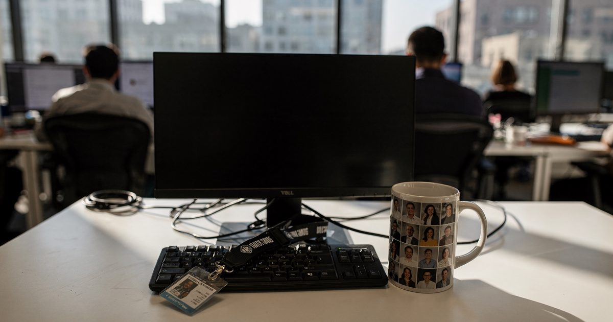 Hard morning sun through floor-to-ceiling glass in a San Francisco open-plan tech office, one unoccupied desk in the foreground with a single coffee mug and a.