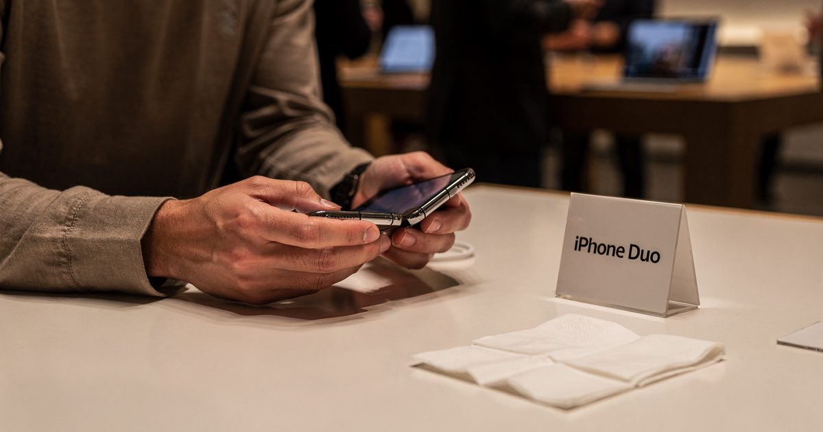 Medium shot under warm evening lights on an Apple Store sales floor, a shopper cropped at the eyes unfolding a new foldable phone on a white table beside a.