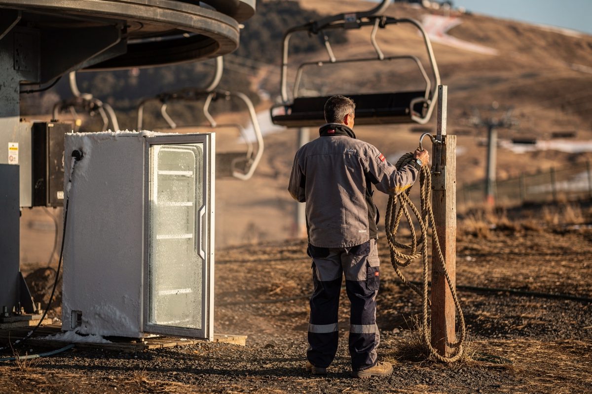 Medium shot at the base of an idle chairlift in late-afternoon light, empty chairs hanging motionless over cracked dry earth while a worker in a resort jacket.