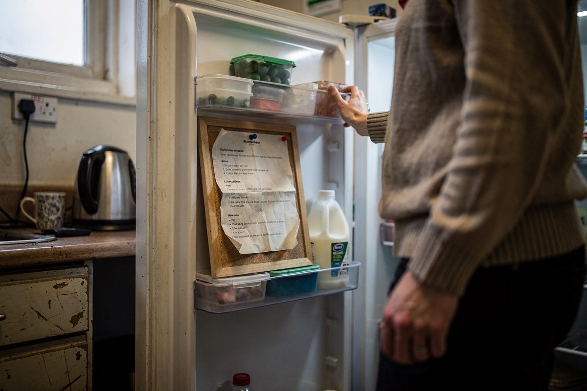 Mid-shot in a cramped university department kitchen lit by morning light from a single high window, a researcher seen only from the shoulders down holding open.