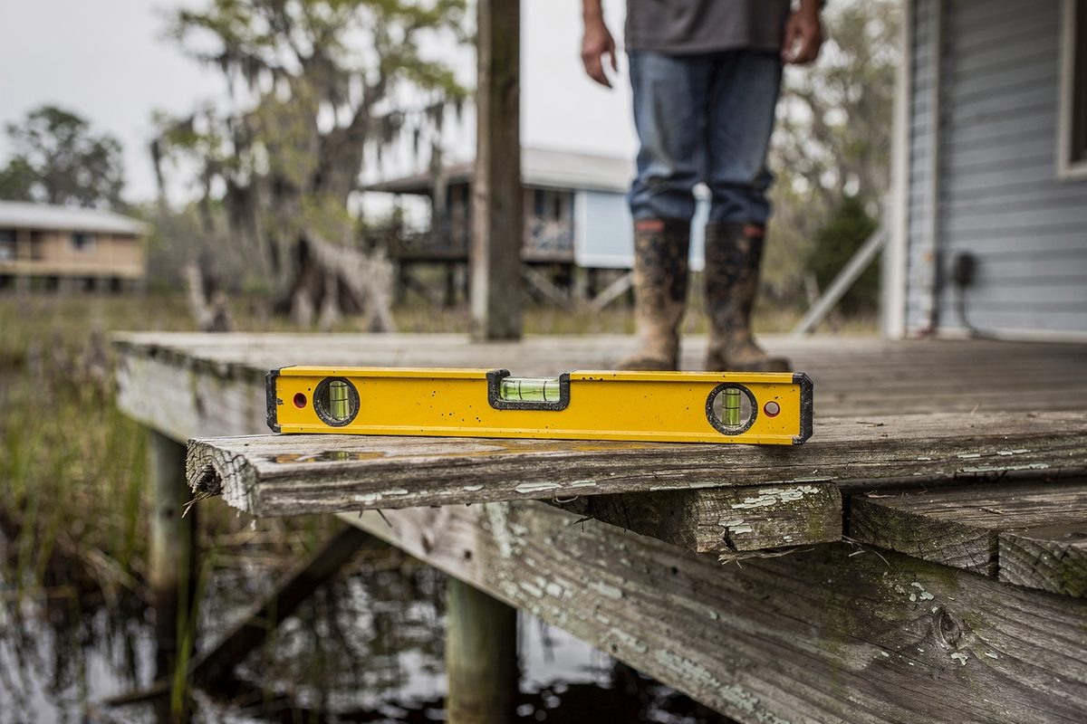 Close detail in flat overcast light of a spirit level resting on the rail of a raised bayou porch with its bubble drifted off center, a homeowner in rubber.