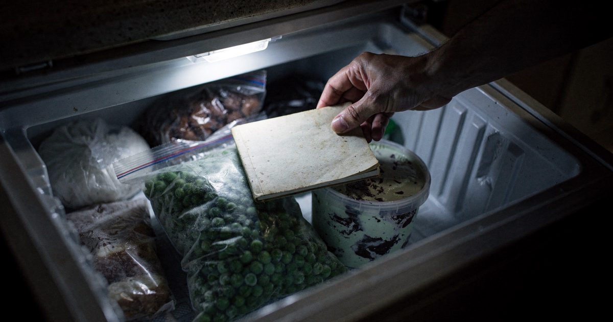 Close detail inside an open household chest freezer in a dark kitchen at night, lit only by the freezer's own interior bulb, a hand cropped at the wrist.