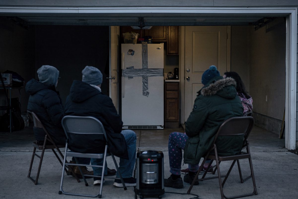 Wide shot from the driveway of a suburban Madison garage in flat early-morning light, a family seen from behind on folding chairs around a space heater, and.
