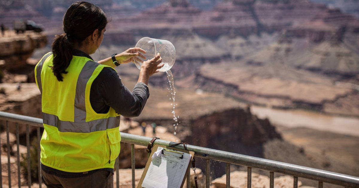 Harsh midday desert light at a canyon-rim overlook as a researcher in a hi-vis vest, seen from behind, tips a graduated glass beaker of water over the.