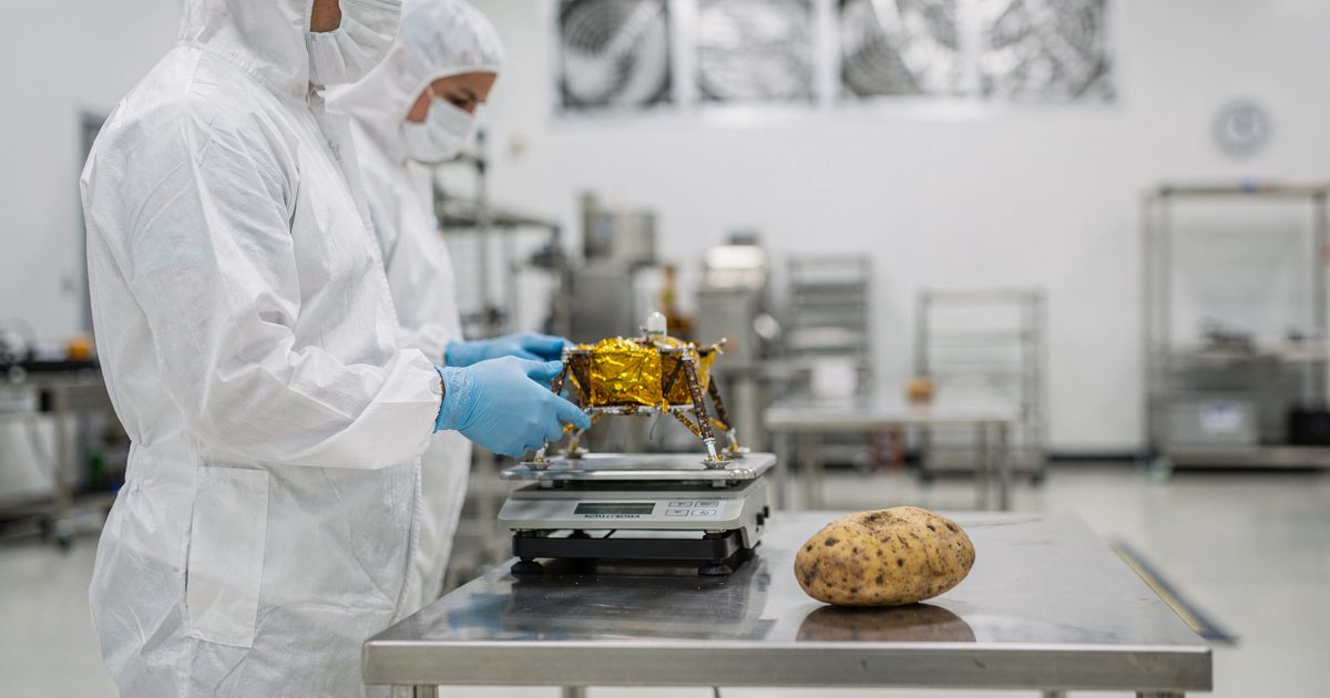 Eye-level medium shot inside a sterile aerospace cleanroom under bright white surgical lighting, two technicians in white bunny suits and face masks seen from.
