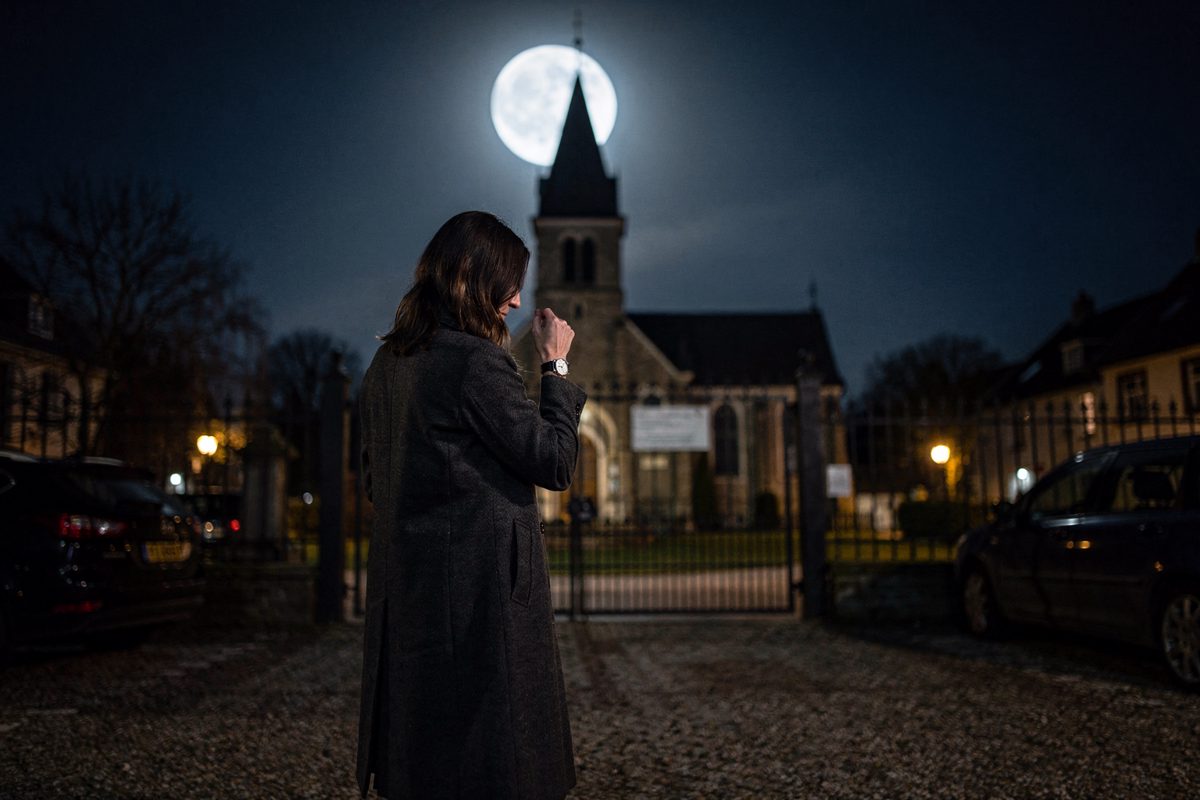 Low-angle night shot from a gravel church car park under a full moon, a woman in a long wool coat seen from the back checking her wristwatch beside a padlocked.