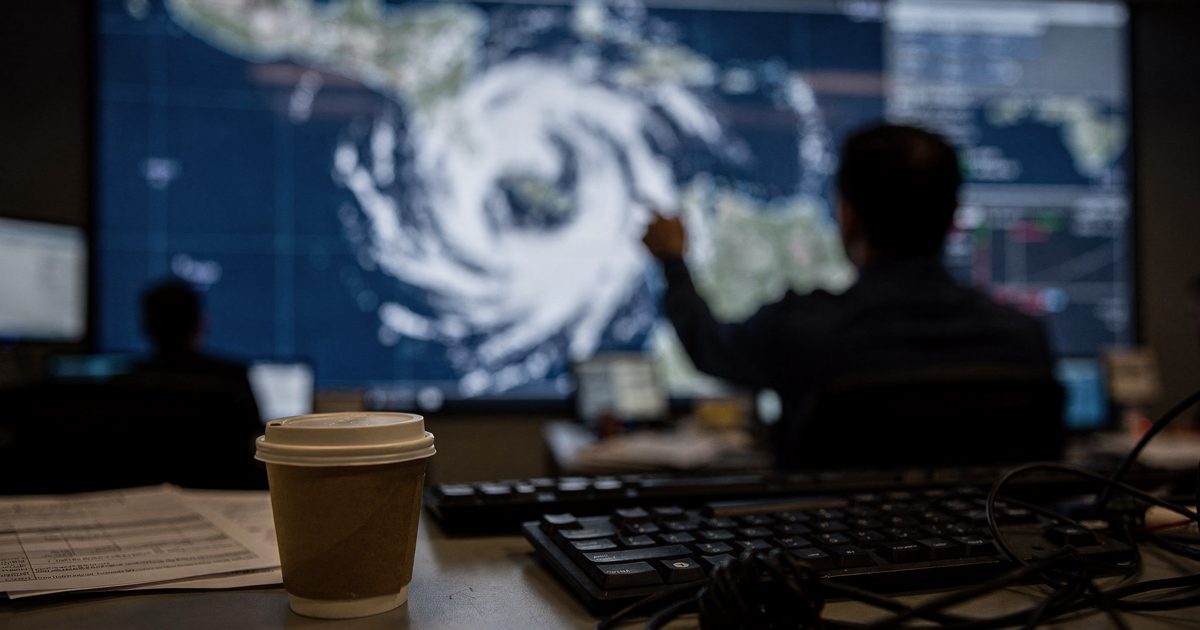 A dark forecast operations room late at night, a part-cropped figure silhouetted against a wall-sized screen glowing with the white spiral of a cyclone over.