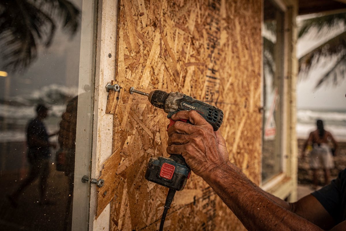 Close-up of weathered hands drilling a sheet of plywood over the window of a beachfront shop in warm late-afternoon Hawaiian light, palm fronds blurring in.