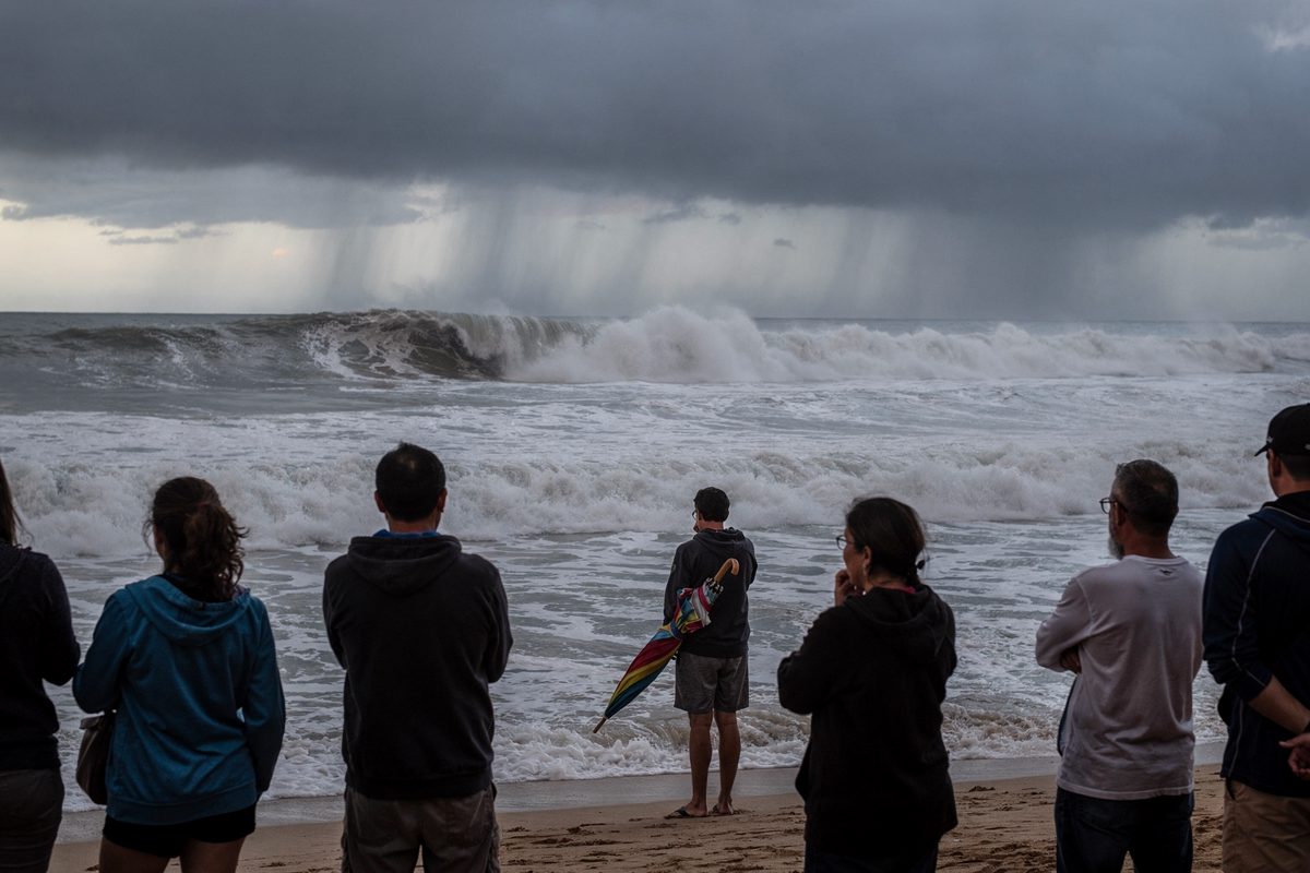 Wide shot from behind a small crowd standing on a grey Hawaiian shoreline at dawn, watching heavy churning surf under low racing clouds, one person clutching a.