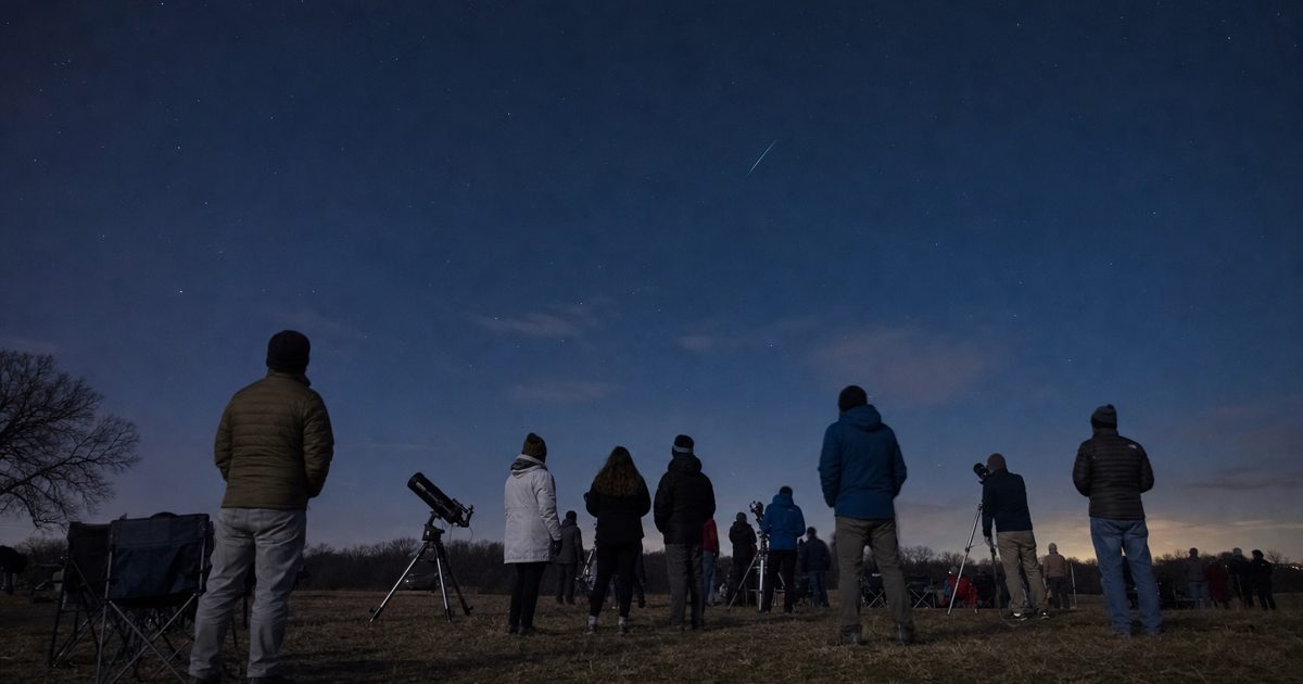 Wide nighttime news photograph of a public astronomy gathering in an open field, anonymous spectators seen from behind beside tripods and telescopes, dark blue.